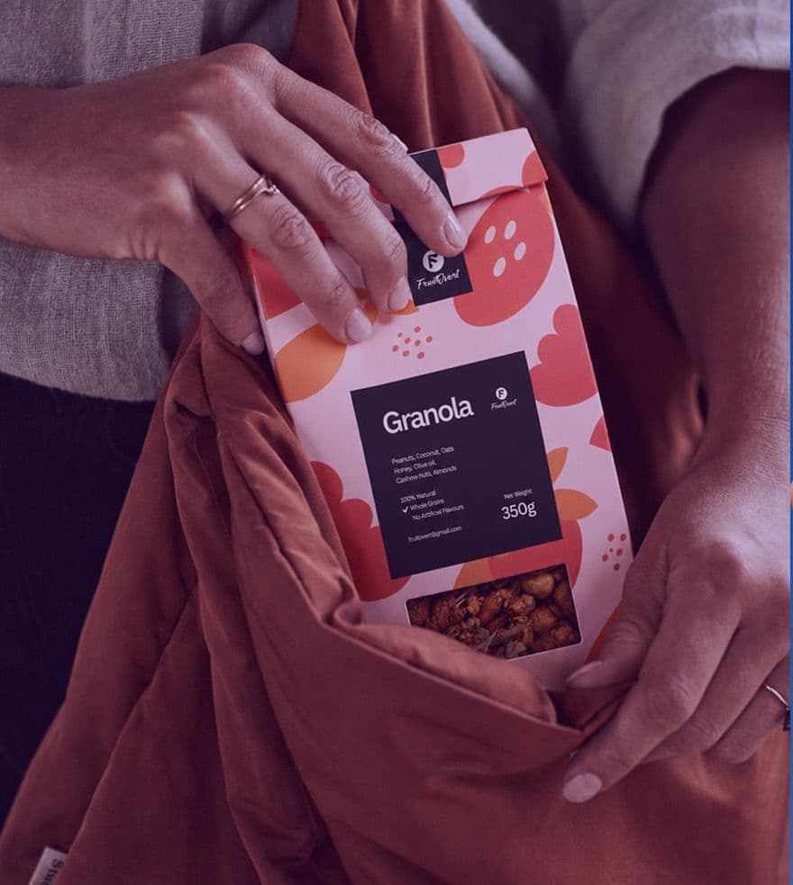 Hands tucking a FruitOvert granola pack into a rust-coloured bag